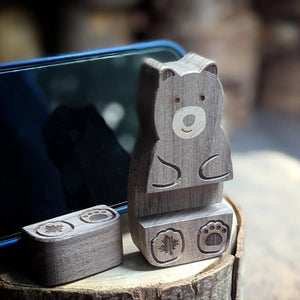 A small carved wooden bear-shaped object stands on a matching rectangular base on a rustic wood slice, with a smartphone reflected behind it in a blurred outdoor setting.