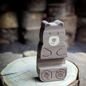 A small carved wooden bear figurine made from layered gray-brown wood, standing on a round wood slice with a blurred rustic background.