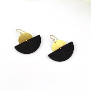 A pair of modern semi-circular drop earrings with a black wood-textured lower half and a gold metal-textured upper half, hanging from gold fishhook wires. The earrings are displayed on a clean white surface with dried pampas grass in the background.