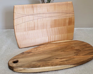 Two wooden serving boards displayed indoors on a light carpet in front of a plain wall, with a small plant in a glass vase behind the taller board.