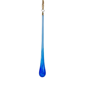 A long, teardrop-shaped glass Christmas ornament in a gradient of blue, hanging from a pine branch against a white background.