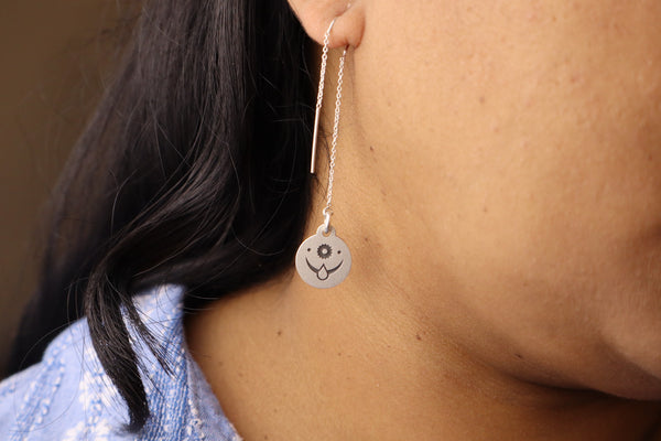 Close-up of a sterling silver threader earring with an engraved celestial face design, worn on a woman’s ear.