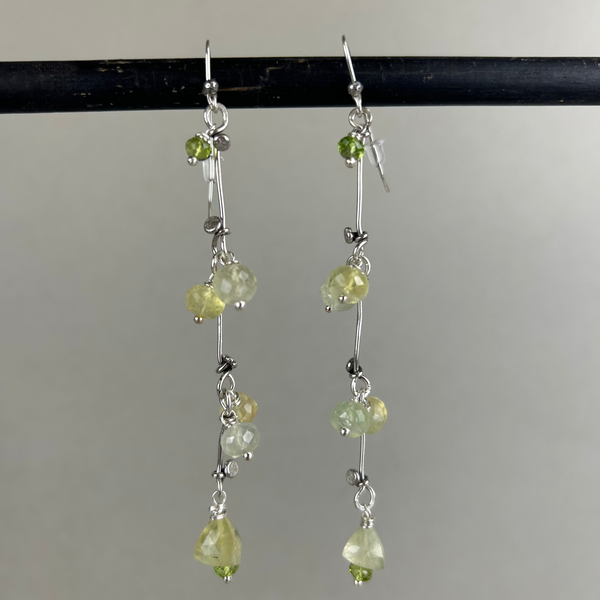 Sterling silver earrings handcrafted in a flowing vine shape with green prehnite and peridot gemstones, laid flat on a light background. Nature-inspired design with a fresh, botanical aesthetic.