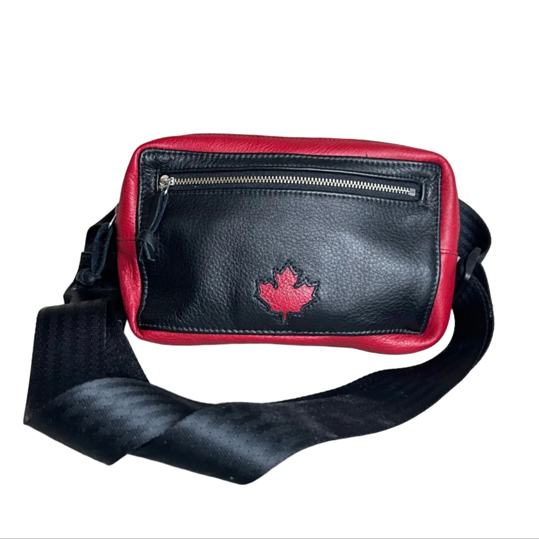 Black and red leather crossbody bag with a red maple leaf logo, featuring a front zippered pocket and a wide black strap, set against a neutral gray background.