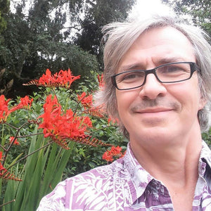 Man with glasses and gray hair standing in front of red flowers and greenery
