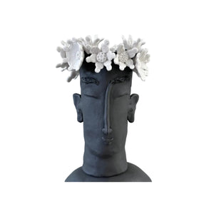 A sculptural black ceramic bust of a face with closed eyes, adorned with white coral-like flowers on the head and a small white flower held near the shoulder, photographed against a plain light gray background.
